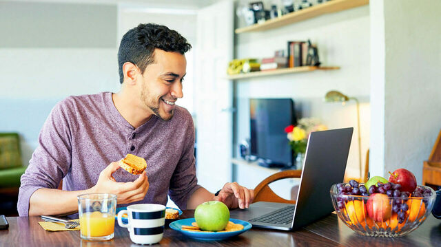 Mann mit Brotscheibe in der Hand sitzt an Computer und lacht