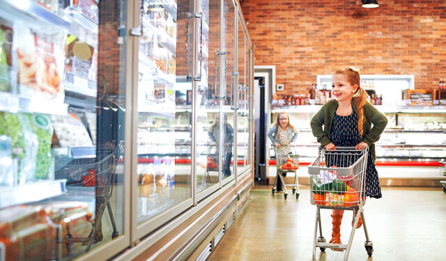 Ein kleines Mädchen mit Einkaufswagen blickt in die Kühlschränke im Supermarkt.