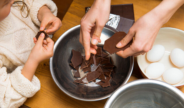 Eine menschliche Hand zerkleinert Schokolade in eine Rührschüssel zum Backen. Ein Kind hilft der Person dabei.